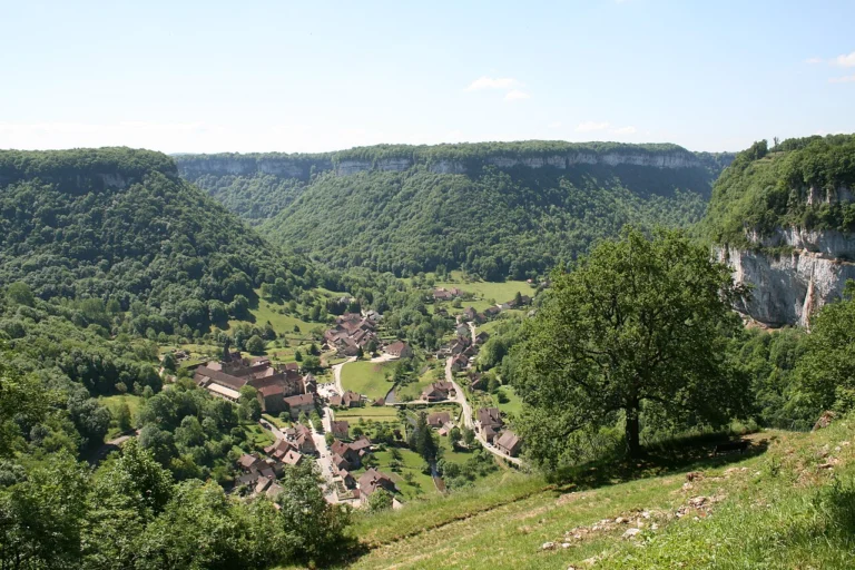 Village de maisons aux toits de tuiles au fond d'une vallée verte encadrée de falaises calcaires boisées.