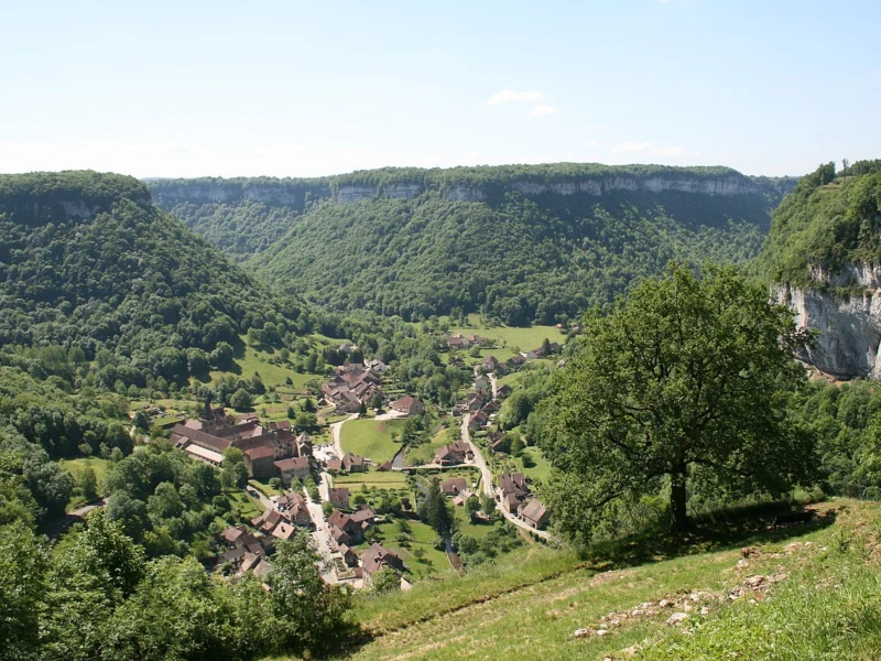 Village de maisons aux toits de tuiles au fond d'une vallée verte encadrée de falaises calcaires boisées.
