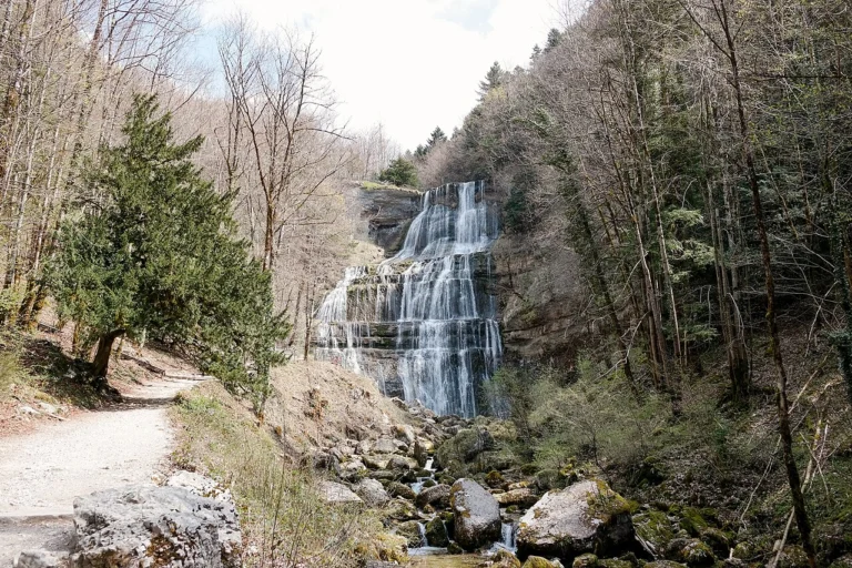 Haute cascade en gradins tombant d'une falaise dans une forêt de printemps, avec un sentier au premier plan.