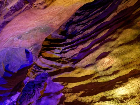 Paroi rocheuse ondulée d'une grotte, éclairée de lumières violettes et dorées.