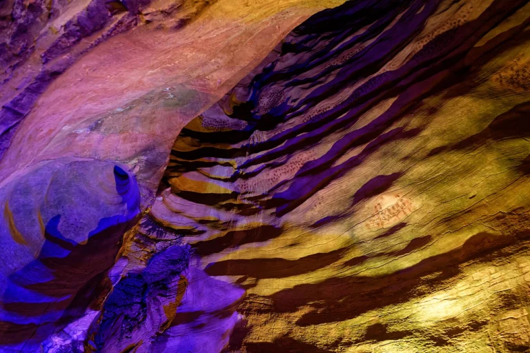 Paroi rocheuse ondulée d'une grotte, éclairée de lumières violettes et dorées.