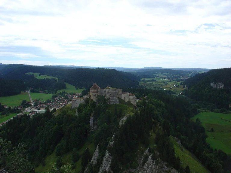 Forteresse sur un sommet rocheux couvert de forêt, dominant une vallée verdoyante et des villages, sous un ciel clair.