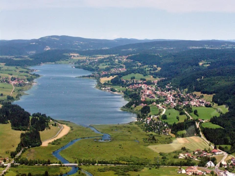 Lac allongé vu du ciel, bordé de prairies, de villages et de collines boisées, avec une rivière sinueuse au premier plan.