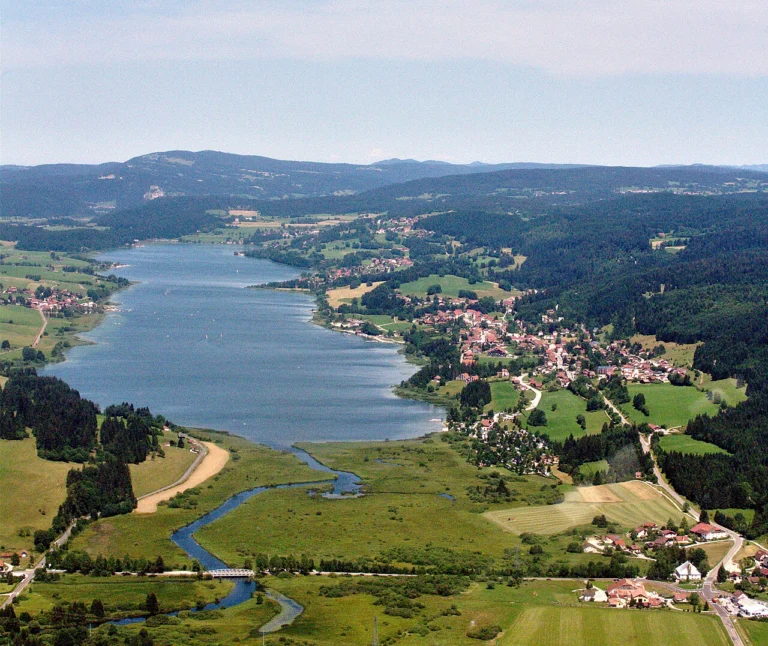 Lac allongé vu du ciel, bordé de prairies, de villages et de collines boisées, avec une rivière sinueuse au premier plan.