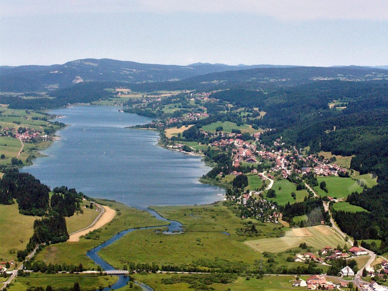 Lac allongé vu du ciel, bordé de prairies, de villages et de collines boisées, avec une rivière sinueuse au premier plan.