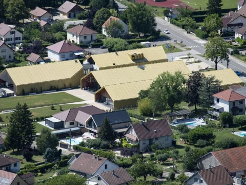 Vue plongeante sur un quartier résidentiel avec, au centre, un grand bâtiment aux toits clairs entouré de verdure.