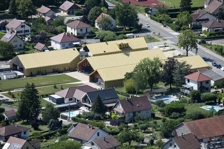 Vue plongeante sur un quartier résidentiel avec, au centre, un grand bâtiment aux toits clairs entouré de verdure.