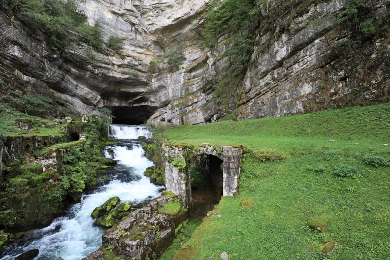 Eau vive jaillissant d'une grande cavité au pied d'une haute falaise calcaire, près d'un mur en pierre et d'une pelouse.