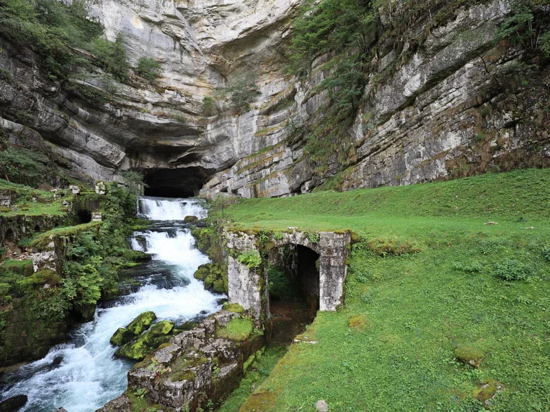 Eau vive jaillissant d'une grande cavité au pied d'une haute falaise calcaire, près d'un mur en pierre et d'une pelouse.