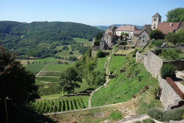 Maisons de pierre et clocher au sommet d'un coteau en terrasses plantées de vignes, sous un ciel bleu.