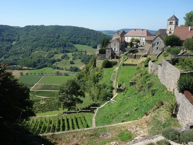 Maisons de pierre et clocher au sommet d'un coteau en terrasses plantées de vignes, sous un ciel bleu.