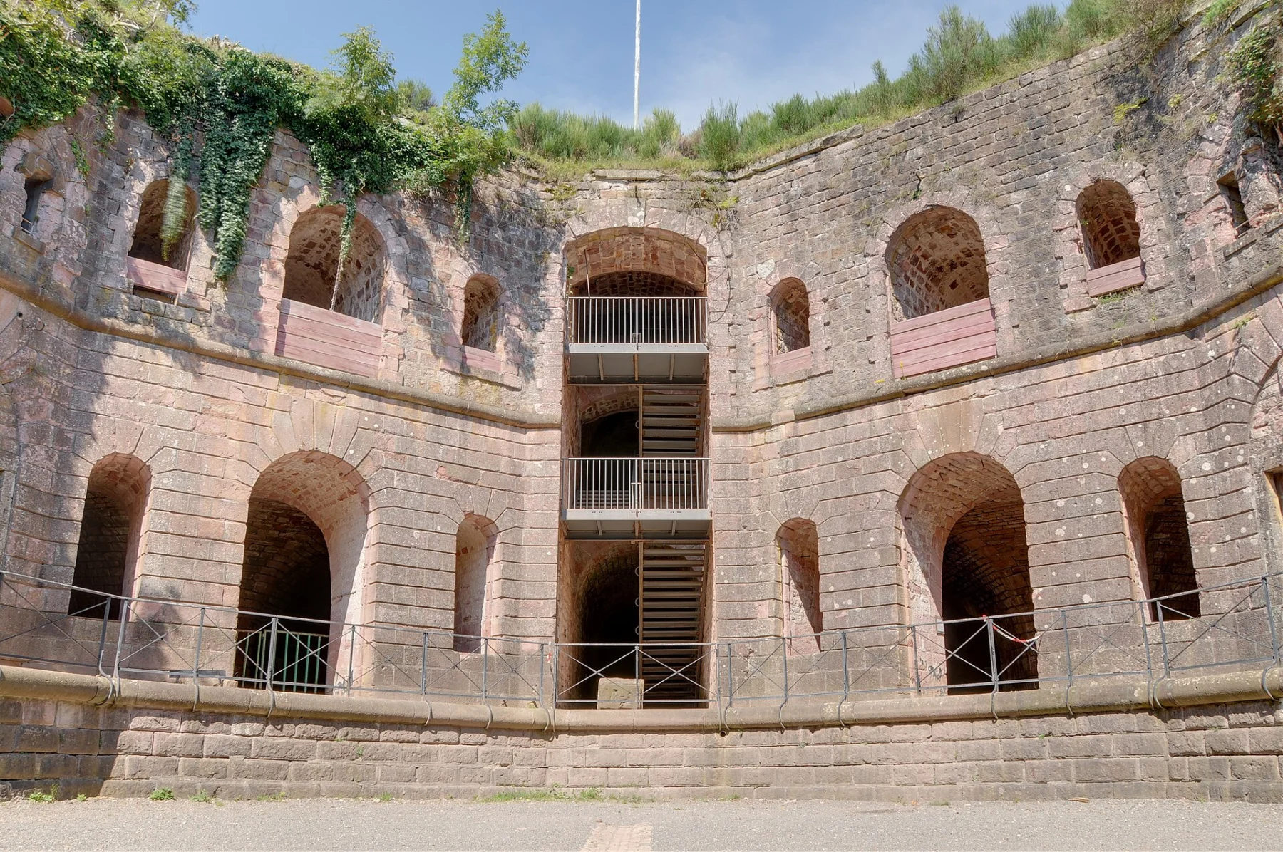 Cour intérieure cernée de hauts murs de pierre percés d’arcades sur plusieurs niveaux, avec un escalier métallique au centre et de la végétation au sommet des murs.
