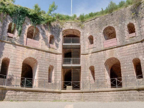 Cour intérieure cernée de hauts murs de pierre percés d’arcades sur plusieurs niveaux, avec un escalier métallique au centre et de la végétation au sommet des murs.