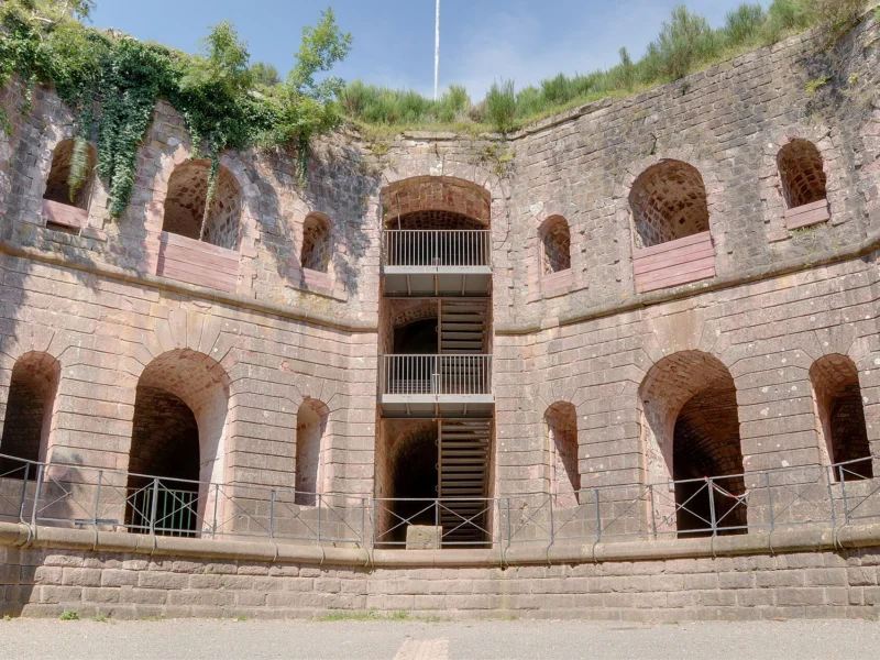 Cour intérieure cernée de hauts murs de pierre percés d’arcades sur plusieurs niveaux, avec un escalier métallique au centre et de la végétation au sommet des murs.