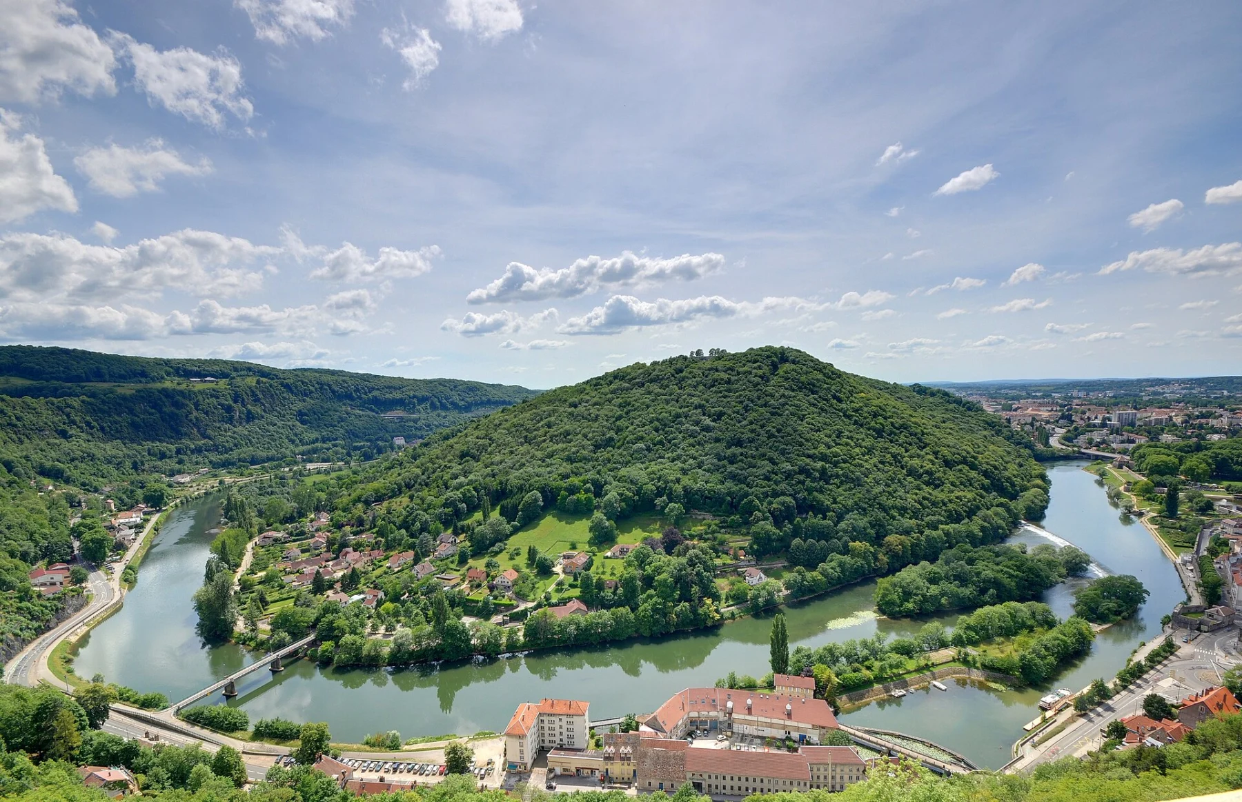 Vue plongeante sur un méandre de rivière vert sombre contournant une colline boisée, avec des bâtiments aux toits rouges au premier plan et la ville au loin.