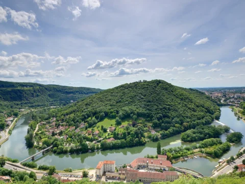 Vue plongeante sur un méandre de rivière vert sombre contournant une colline boisée, avec des bâtiments aux toits rouges au premier plan et la ville au loin.