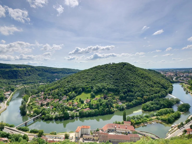 Vue plongeante sur un méandre de rivière vert sombre contournant une colline boisée, avec des bâtiments aux toits rouges au premier plan et la ville au loin.