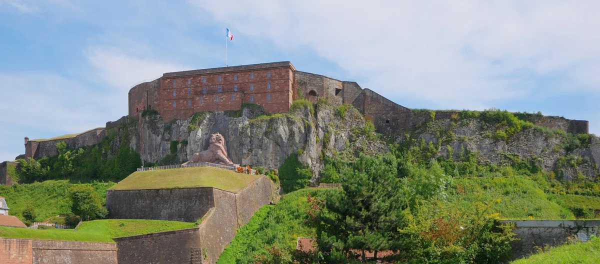 Caserne en pierre rose au sommet d’un rocher, remparts herbeux en contrebas et lion sculpté sur une terrasse, sous un ciel bleu.