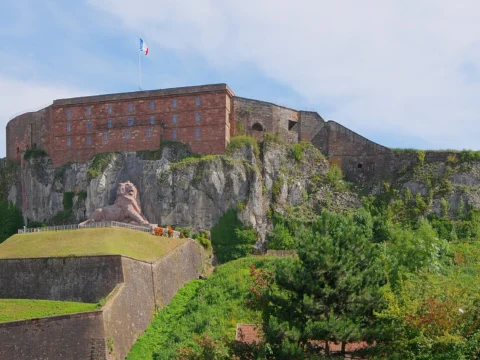 Massive caserne de grès rose au sommet d’une falaise calcaire, remparts gazonnés au premier plan et lion sculpté sur la gauche, sous un ciel bleu.