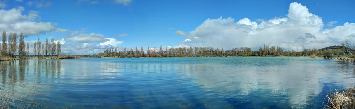 Vaste plan d’eau bleu bordé de peupliers et de rives boisées, sous un ciel traversé de gros nuages blancs.