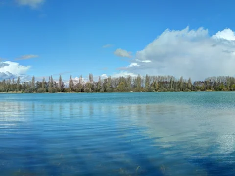 Vaste plan d’eau bleu bordé de peupliers et de rives boisées, sous un ciel traversé de gros nuages blancs.