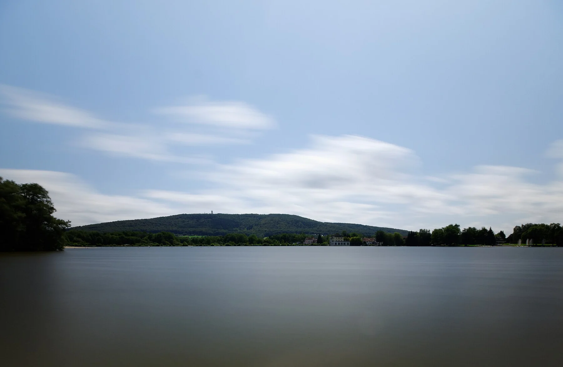 Plan d’eau lisse aux reflets gris sous un ciel traversé de nuages étirés, bordé d’une ligne de forêt et d’une colline boisée à l’horizon.
