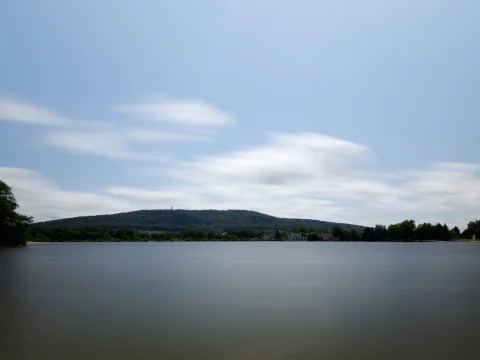 Plan d’eau lisse aux reflets gris sous un ciel traversé de nuages étirés, bordé d’une ligne de forêt et d’une colline boisée à l’horizon.