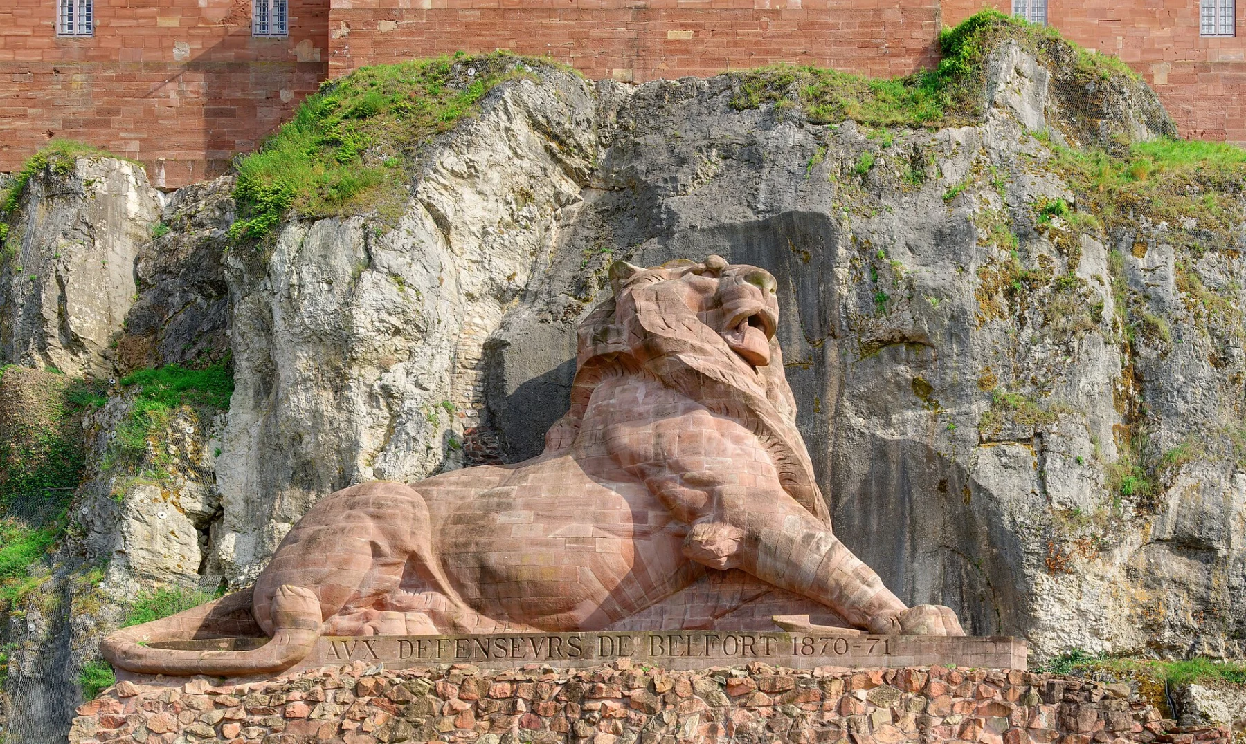 Grand lion couché sculpté en grès rose, tête relevée, adossé à une falaise calcaire grise tapissée de végétation, sous les murs de brique de la citadelle.