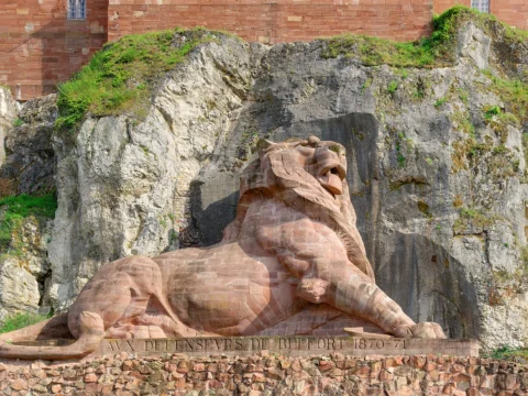 Grand lion couché sculpté en grès rose, tête relevée, adossé à une falaise calcaire grise tapissée de végétation, sous les murs de brique de la citadelle.