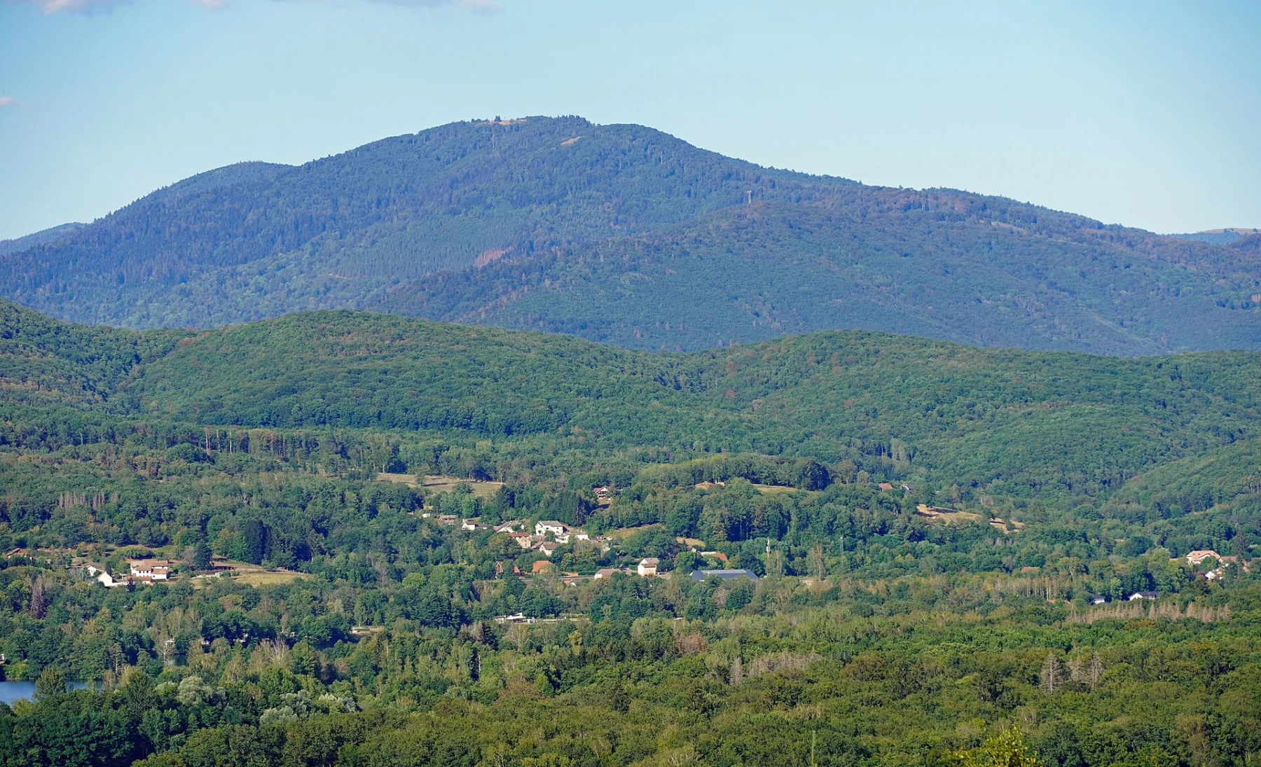 Sommet boisé arrondi se détachant sur un ciel pâle, au-dessus de collines couvertes de forêt et de quelques maisons dans la vallée.