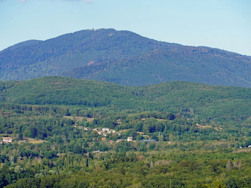 Sommet boisé arrondi se détachant sur un ciel pâle, au-dessus de collines couvertes de forêt et de quelques maisons dans la vallée.