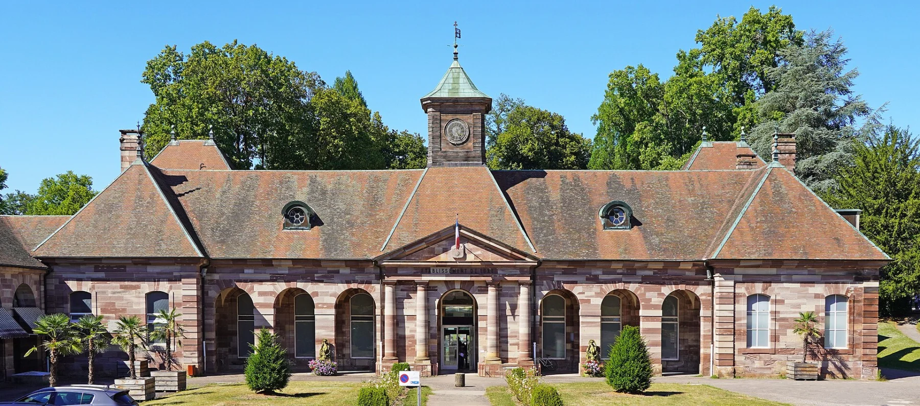 Long bâtiment de grès rose à arcades, coiffé d’un toit à lucarnes et d’un petit clocheton central, devant une pelouse plantée et de grands arbres.