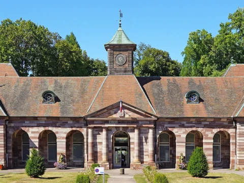 Long bâtiment de grès rose à arcades, coiffé d’un toit à lucarnes et d’un petit clocheton central, devant une pelouse plantée et de grands arbres.