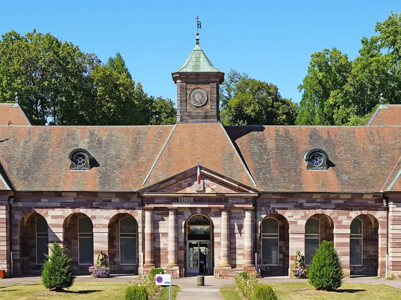 Long bâtiment de grès rose à arcades, coiffé d’un toit à lucarnes et d’un petit clocheton central, devant une pelouse plantée et de grands arbres.