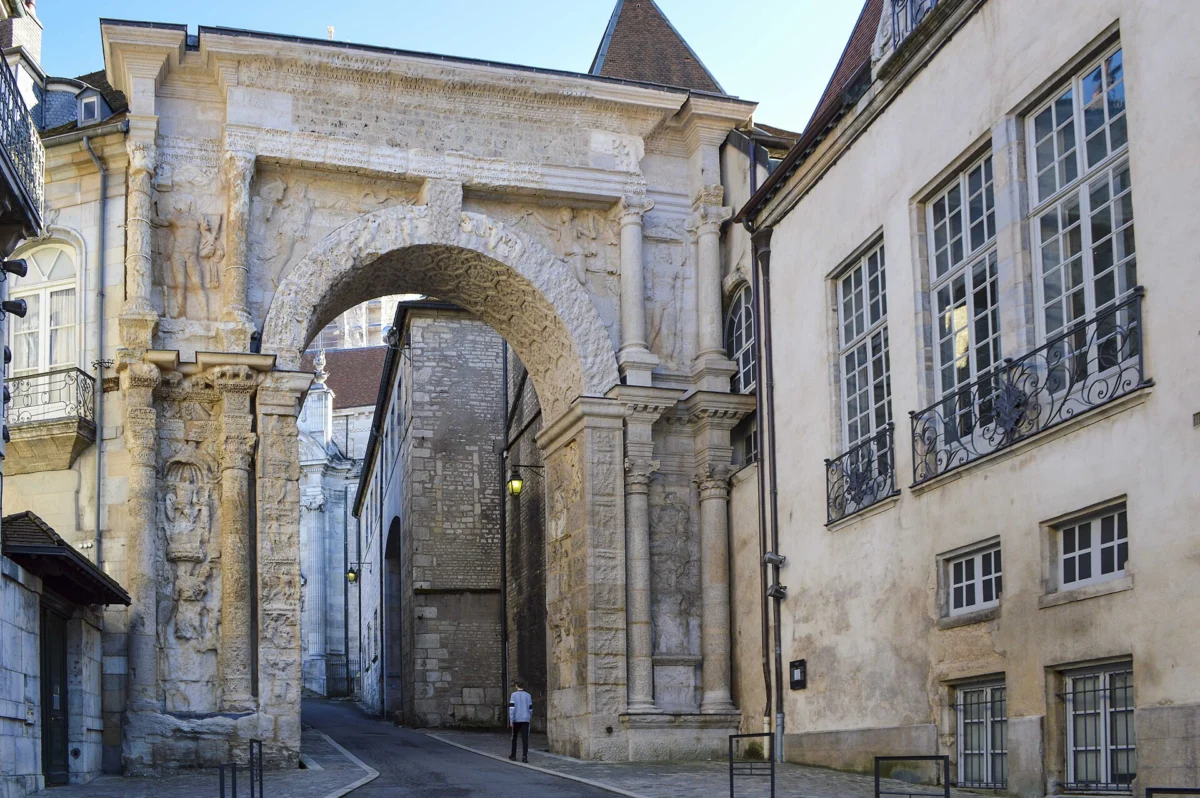 Arc de triomphe antique en pierre sculptée, encadré de façades anciennes, ouvrant sur une rue pavée.