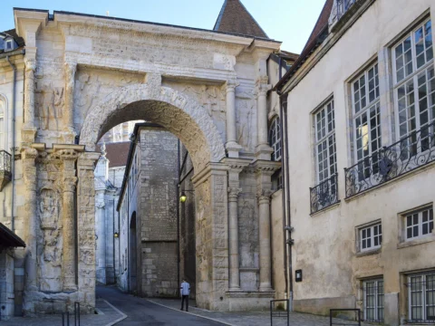 Arc de triomphe antique en pierre sculptée, encadré de façades anciennes, ouvrant sur une rue pavée.