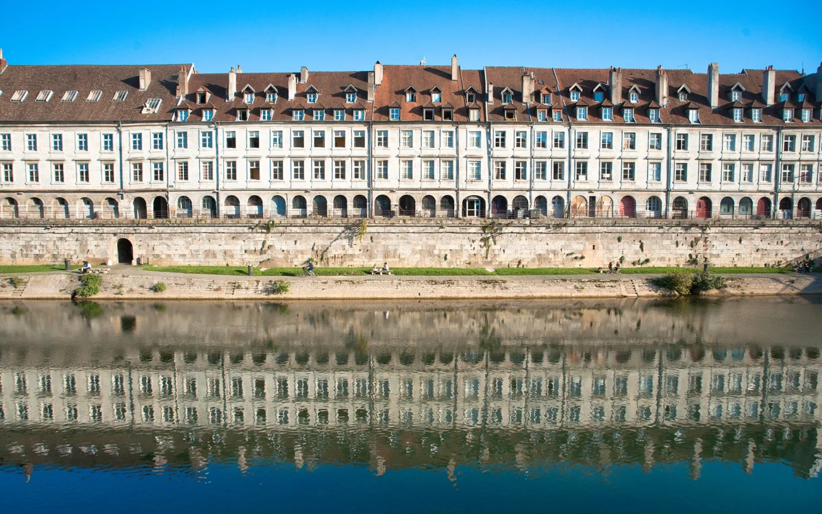 Longue façade classique à arcades et lucarnes au bord d’un quai de pierre, reflétée dans l’eau calme de la rivière sous un ciel bleu.