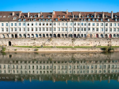Longue façade classique à arcades et lucarnes au bord d’un quai de pierre, reflétée dans l’eau calme de la rivière sous un ciel bleu.