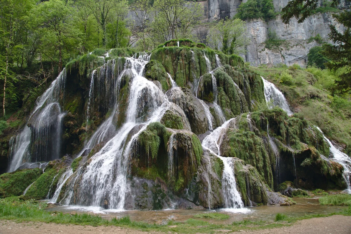 Nombreux filets d’eau blanche dévalant des gradins de tuf couverts de mousse verte, au pied d’une falaise et d’arbres.