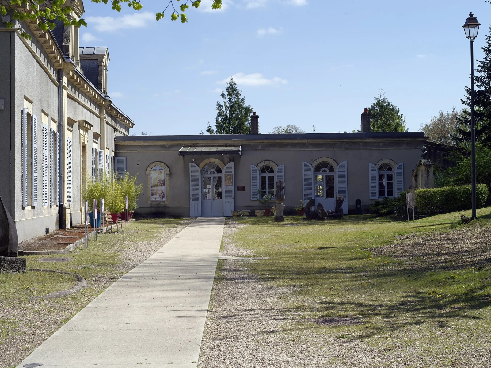 Allée de gravier menant à un pavillon classique aux volets clairs, encadrée d’une aile à hautes fenêtres et d’un lampadaire, sous un ciel bleu.