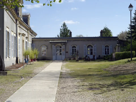 Allée de gravier menant à un pavillon classique aux volets clairs, encadrée d’une aile à hautes fenêtres et d’un lampadaire, sous un ciel bleu.