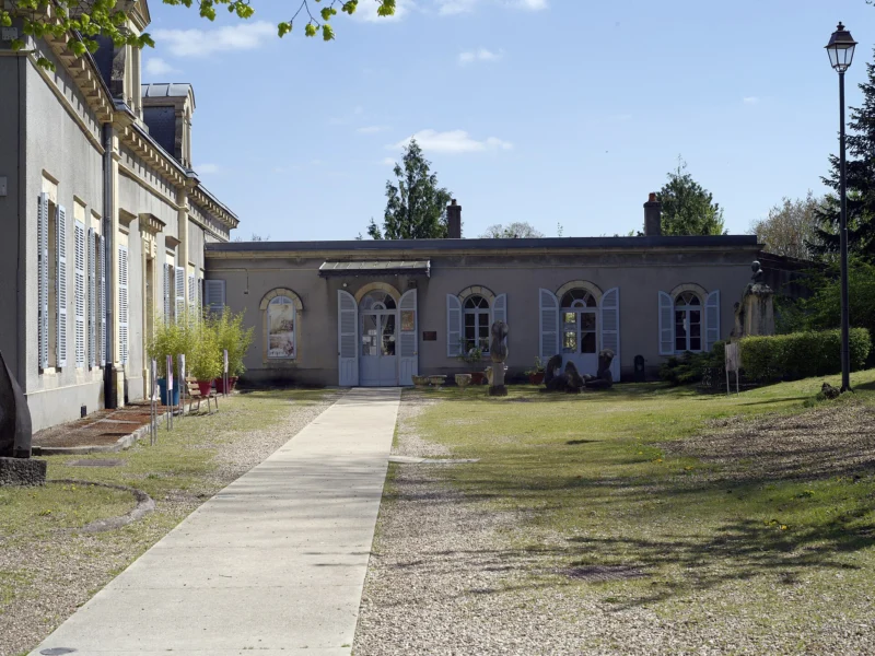 Allée de gravier menant à un pavillon classique aux volets clairs, encadrée d’une aile à hautes fenêtres et d’un lampadaire, sous un ciel bleu.