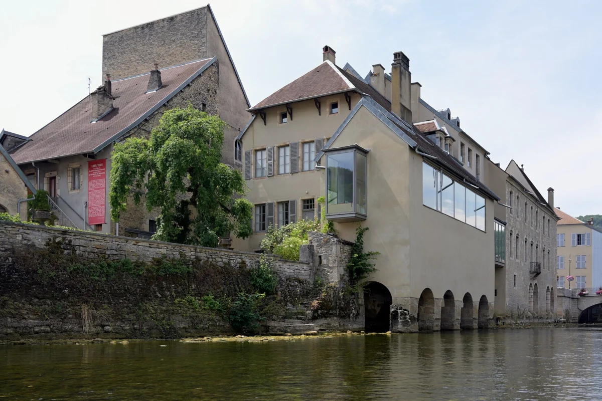 Bâtiments anciens aux façades claires, dont une avancée vitrée, posés sur un mur de pierre et des arches au bord de la rivière.