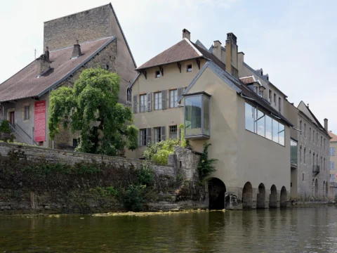 Bâtiments anciens aux façades claires, dont une avancée vitrée, posés sur un mur de pierre et des arches au bord de la rivière.