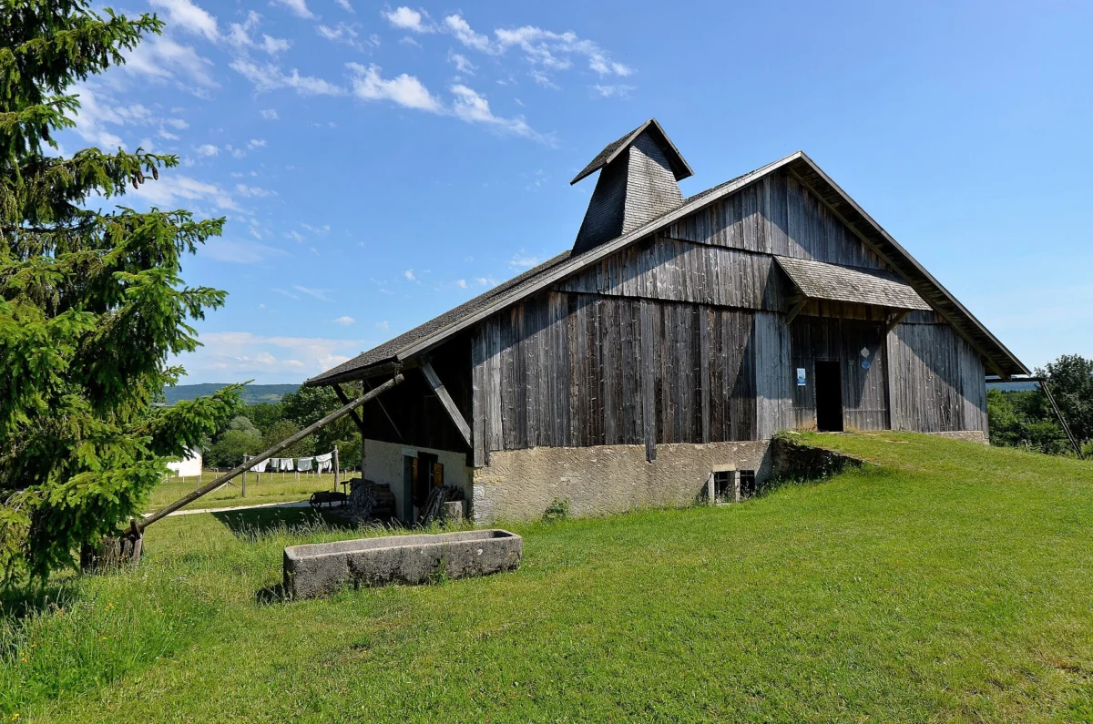 Ferme au grand toit gris vue de trois quarts, bardée de planches, avec une cheminée pyramidale en bois coiffée d’un petit toit et un abreuvoir de pierre dans l’herbe.