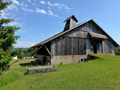 Ferme au grand toit gris vue de trois quarts, bardée de planches, avec une cheminée pyramidale en bois coiffée d’un petit toit et un abreuvoir de pierre dans l’herbe.