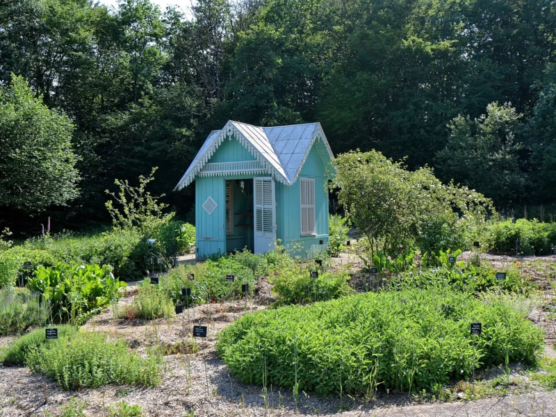 Petit kiosque de bois peint en bleu au milieu de massifs de plantes aromatiques et médicinales, sous des arbres.