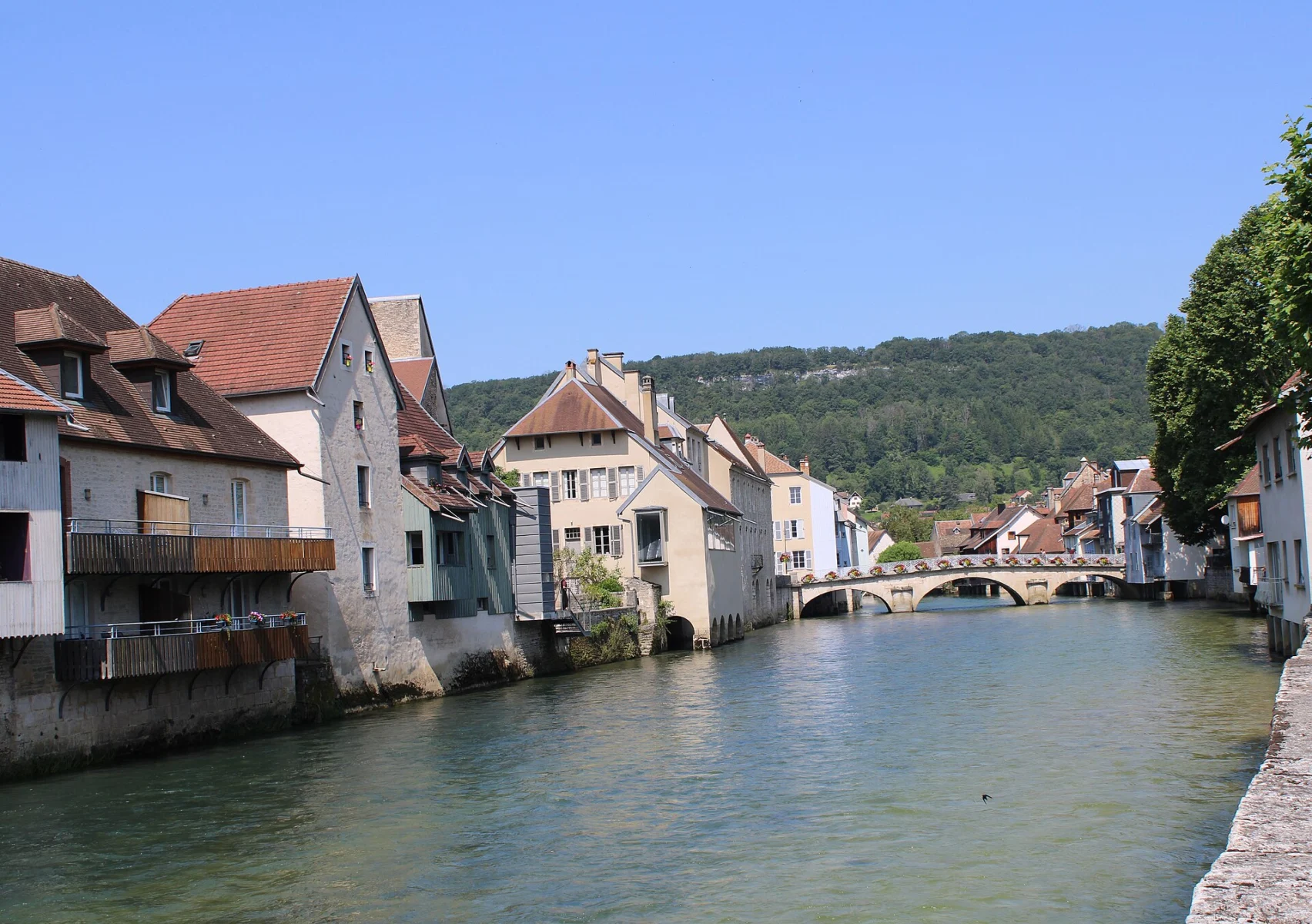 Rivière calme bordée de maisons anciennes aux toits de tuiles, un pont de pierre à arches au fond et une colline boisée sous un ciel bleu.