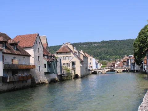 Rivière calme bordée de maisons anciennes aux toits de tuiles, un pont de pierre à arches au fond et une colline boisée sous un ciel bleu.