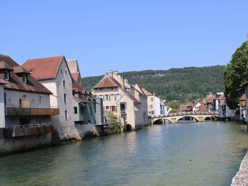 Rivière calme bordée de maisons anciennes aux toits de tuiles, un pont de pierre à arches au fond et une colline boisée sous un ciel bleu.